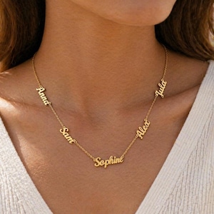 May include: A gold-colored necklace with multiple name pendants. The names "Anna", "Sam", "Sophie", "Alex", and "Julia" are displayed in a delicate, cursive font. The necklace is worn against a cream-colored top.