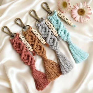 May include: Macrame keychains in shades of dusty rose, brown, gray, and teal, each with a tassel and a wooden bead strand spelling "MAMA." The keychains have bronze-colored clasps and are arranged on a white satin surface.