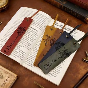Può includere: Una collezione di segnalibri in pelle di vari colori, tra cui rosso, giallo, blu navy e verde, ognuno con un design floreale e il nome "Olivia". I segnalibri sono posizionati su un libro aperto, con un cordino in pelle.