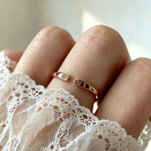 May include: A rose gold open ring featuring a small engraved heart symbol on one end and the letter C on the other end. The band is smooth and polished, designed with a gap between the two ends, resting on a finger against white lace fabric.