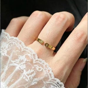 Könnte beinhalten: Ein goldfarbener offener Ring mit zwei flachen, abgerundeten Enden. Jedes Ende ist mit einem einzelnen Großbuchstaben graviert, konkret den Buchstaben F und S, die sich über die offene Lücke des Rings hinweg gegenüberstehen.