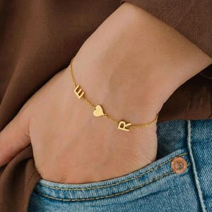 Op de afbeelding: Een goudkleurige armband met de letters "E", een hartje en "R" aan een fijne ketting. De armband wordt om een pols gedragen, met een bruin overhemd en een blauwe jeans op de achtergrond.