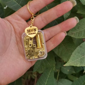 Può includere: Un portachiavi carillon dorato con una custodia in plastica trasparente. Il portachiavi ha un ciondolo a forma di cuore con le iniziali "H S" e la data "04.08.2026". Il meccanismo del carillon è visibile all'interno della custodia trasparente.