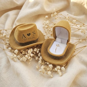 May include: Two miniature cowboy hat ring boxes. One closed hat has the initials "A S" with a heart and the date "01.16.26". The other hat is open, revealing a gold ring with a heart-shaped stone. Both hats are tan.