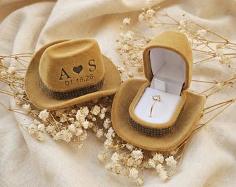 May include: Two miniature cowboy hat ring boxes. One closed hat has the initials "A S" with a heart and the date "01.16.26". The other hat is open, revealing a gold ring with a heart-shaped stone. Both hats are tan.