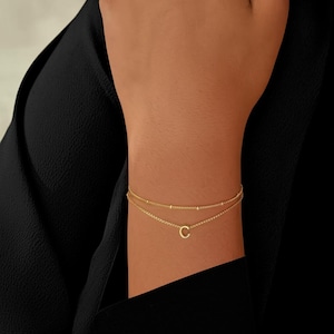 Könnte beinhalten: Ein goldfarbenes, mehrschichtiges Armband mit filigranem Kettendesign. Die obere Schicht hat kleine, gleichmäßig verteilte Perlen, während die untere Schicht einen Anhänger mit dem Buchstaben "C" hat. Das Armband wird am Handgelenk getragen.