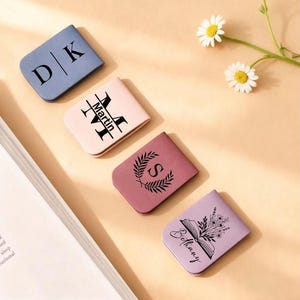 Può includere: Quattro segnalibri magnetici personalizzati in vari colori: blu, rosa, malva e lavanda. Ogni segnalibro presenta un design unico, tra cui iniziali, un nome, una corona d'alloro con una lettera e un'illustrazione floreale con un nome. Le margherite sono sullo sfondo.