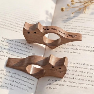 Può includere: Due fermapagina in legno a forma di gatto con la scritta "Just one more page..." incisa. I fermapagina sono marroni e hanno un foro per il dito. Un gatto ha un semplice volto scolpito.