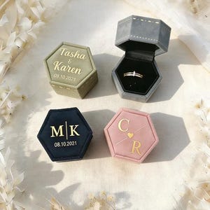 Könnte beinhalten: Vier sechseckige Ringboxen in verschiedenen Farben: Olivgrün, Grau, Marineblau und Rosa. Die olivgrüne Box trägt die Namen "Tasha & Karen" und das Datum "08.10.2021" in goldfarbenen Buchstaben. Die graue Box ist geöffnet und zeigt einen Ring. Die marineblaue Box hat "MK" und das Datum. Die rosa Box hat "C R".