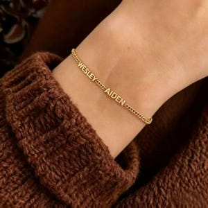 Puede incluir: Una pulsera de cadena dorada con los nombres "WESLEY" y "AIDEN". La pulsera se lleva en la muñeca, sobre un fondo de jersey marrón. La pulsera es de oro.