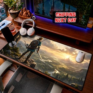 May include: A rectangular mouse mat with a landscape scene and a figure with a sword and bow. The mat has a detailed illustration of a character on a cliff overlooking a valley. A white mouse and headphones are also visible.