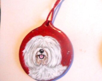 May include: A red, teardrop-shaped ornament featuring a hand-painted portrait of a fluffy white dog with a black nose and open mouth. The dog's face is centered on the ornament, which has a red string for hanging.