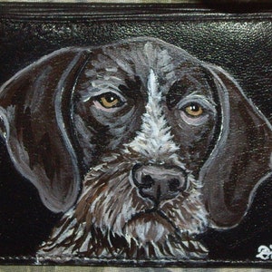 May include: Black leather wallet featuring a hand-painted portrait of a German Wirehaired Pointer. The dog's face is detailed with brown, white, and tan fur, and it has brown eyes. The artist's signature is in the bottom right corner.