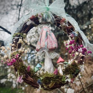 May include: A decorative wreath with a woven base, decorated with moss, miniature mushrooms, and flowers in pink, red, and blue. A large mushroom is in the center, and the wreath is suspended by a sheer ribbon.