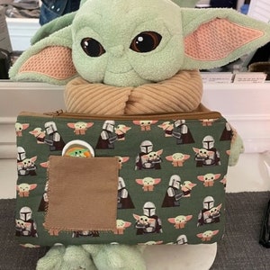 May include: A plush green creature with large dark eyes and pointed ears, wearing a tan scarf, holding a rectangular green zippered pouch. The pouch features a repeating pattern of the creature and a silver-helmeted figure, with a brown pocket.