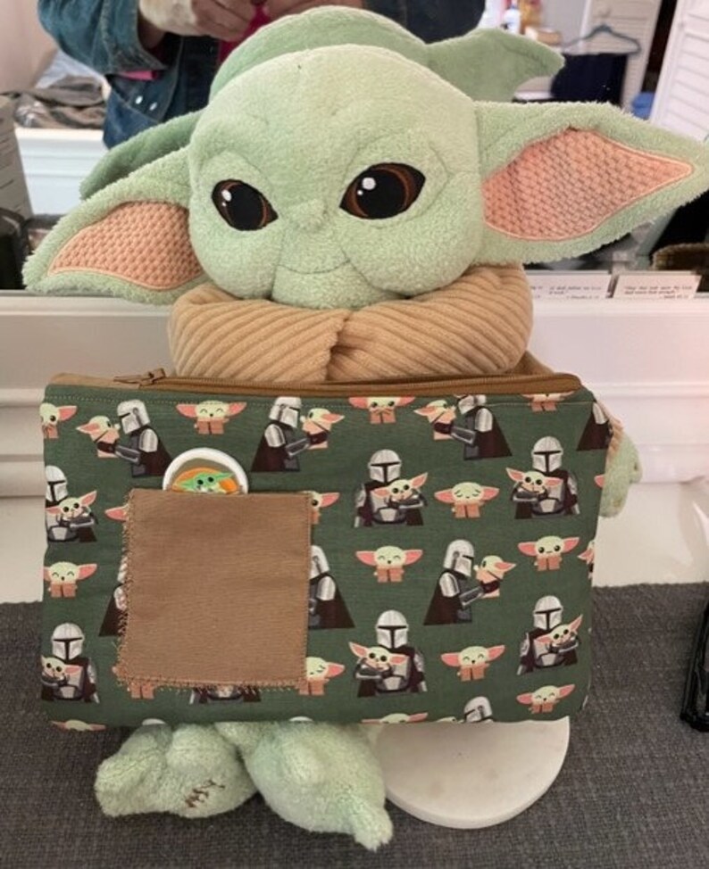 May include: A plush green creature with large dark eyes and pointed ears, wearing a tan scarf, holding a rectangular green zippered pouch. The pouch features a repeating pattern of the creature and a silver-helmeted figure, with a brown pocket.