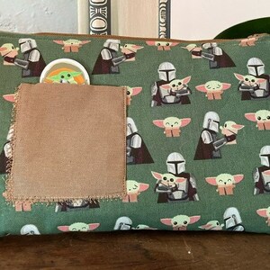 May include: A rectangular green zippered pouch featuring a repeating pattern of a silver-armored character holding a small green creature with large ears. A plain brown fabric square is stitched onto the front, partially obscuring a small circular icon.