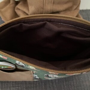 May include: A green zippered pouch featuring a repeating character print, a brown exterior pocket, and a dark brown interior lining. The pouch has a brown zipper closure along the top edge and a small circular button attached to the front pocket.