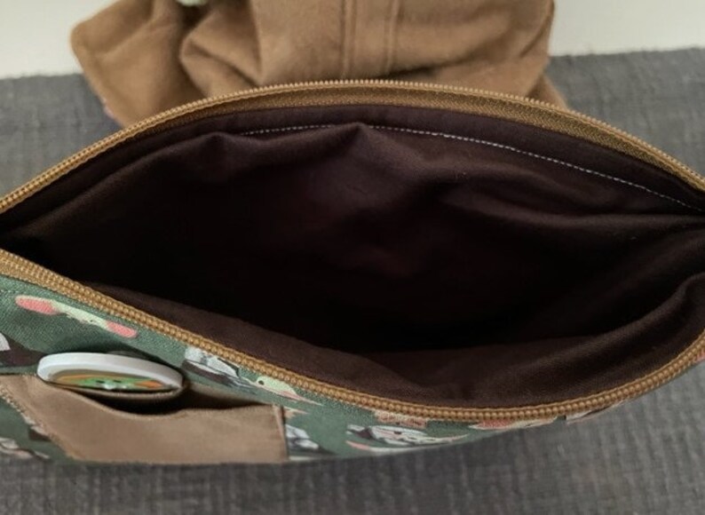 May include: A green zippered pouch featuring a repeating character print, a brown exterior pocket, and a dark brown interior lining. The pouch has a brown zipper closure along the top edge and a small circular button attached to the front pocket.