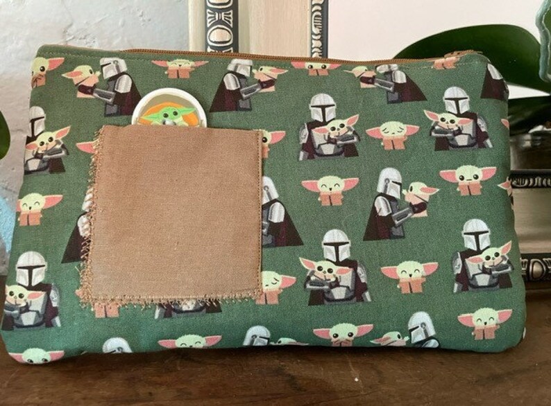 May include: A rectangular green zippered pouch featuring a repeating pattern of a silver-armored character holding a small green creature with large ears. A plain brown fabric square is stitched onto the front, partially obscuring a small circular icon.