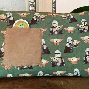 May include: A rectangular green zippered pouch featuring a repeating pattern of a silver-armored character holding a small green creature with large ears. A plain brown fabric square is stitched onto the front, partially obscuring a small circular icon.