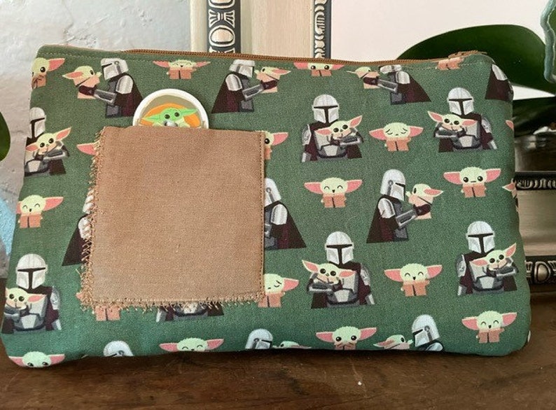 May include: A rectangular green zippered pouch featuring a repeating pattern of a silver-armored character holding a small green creature with large ears. A plain brown fabric square is stitched onto the front, partially obscuring a small circular icon.