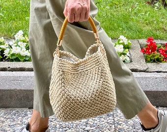 May include: A beige crocheted tote bag with a brown leather handle and gold hardware. The bag is being held by a person wearing khaki pants and white sneakers.