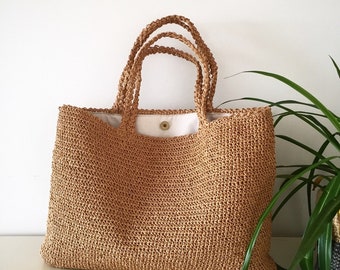 May include: A tan crocheted tote bag with a white fabric lining and a single button closure. The bag has a wide, flat base and two thick, braided handles.