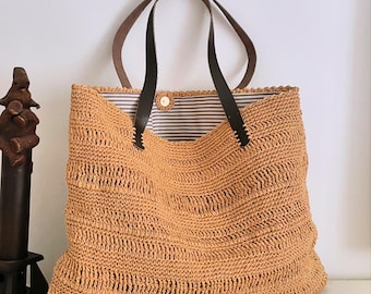 May include: A woven straw tote bag with brown leather handles. The bag has a striped fabric lining and a button closure. The bag is a light tan color and appears to be handmade. The bag is sitting on a white surface.