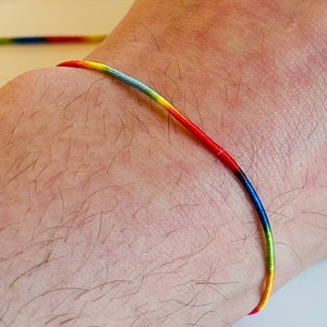May include: A rainbow colored friendship bracelet made of thin string. The bracelet is tied around a person's wrist.