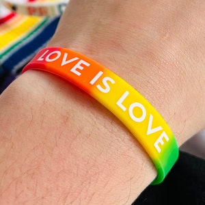 Op de afbeelding: Een regenboogkleurig siliconen armbandje met de tekst "LOVE IS LOVE" in witte letters.