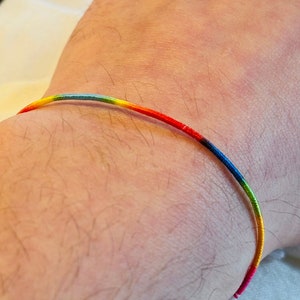 Pode incluir: Um bracelete de corda arco-íris no pulso de uma pessoa. O bracelete é feito de corda fina e está amarrado em um nó.