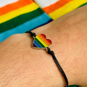 May include: A black string bracelet with a rainbow heart charm. The bracelet is on a person's wrist.