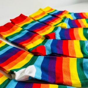 Può includere: Un set di calzini a righe arcobaleno con polsini bianchi. I calzini sono piegati e disposti in fila su una superficie bianca.