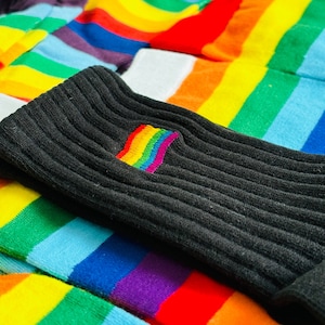 May include: A stack of rainbow striped socks with a black sock on top. The black sock has a rainbow embroidered patch on the side.