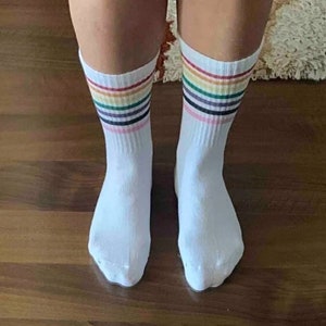 Op de afbeelding: Een paar witte sokken met een regenboogstreepjespatroon rond de enkel.