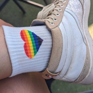 Può includere: Calzini bianchi con un design a forma di cuore arcobaleno. I calzini sono indossati sul piede di una persona con una scarpa bianca.