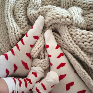 Könnte beinhalten: Weiße Socken mit roten Herzmustern sind zu sehen, getragen von mehreren Personen. Die Socken liegen auf einer beigefarbenen, grob gestrickten Decke. Die Decke hat ein Zopfmuster. Das Bild vermittelt ein gemütliches, familienorientiertes Thema.