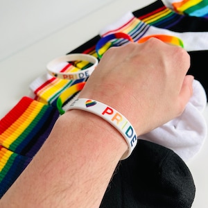 Könnte beinhalten: Ein weißes Silikonarmband mit dem Wort "PRIDE" in Regenbogenfarben und einem kleinen Regenbogenherz. Das Armband wird am Handgelenk getragen.