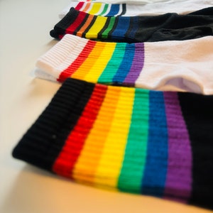 Peut inclure: Quatre paires de chaussettes rayées arc-en-ciel. Les chaussettes sont blanches avec une bande noire en haut et une bande arc-en-ciel en bas. La bande arc-en-ciel est rouge, orange, jaune, verte, bleue, indigo et violette.