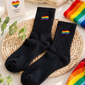 Può includere: Un paio di calzini neri a coste con una piccola bandiera arcobaleno ricamata sul bordo superiore. I calzini sono presentati accanto a un cartoncino bianco con un cuore arcobaleno e la scritta LOVE IS LOVE, e un pezzo di tessuto a righe.