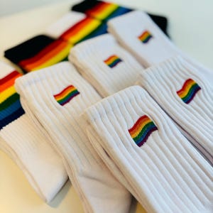 May include: A stack of white socks with rainbow embroidered flags. The socks are folded and arranged in a row.