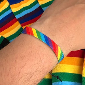 Peut inclure: Un bracelet en tissu tissé présentant un motif de rayures arc-en-ciel diagonales en rouge, orange, jaune, vert, bleu et violet. Le bracelet est fixé par un cordon noir fin et un nœud coulissant, posé sur un tissu à rayures arc-en-ciel assorti.