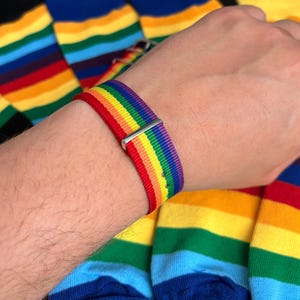 Op de afbeelding: Een regenboog gestreepte horlogeband met een zilverkleurige sluiting wordt getoond op een pols. De kleuren van de band zijn rood, oranje, geel, groen, blauw en paars. Regenboog gestreepte sokken zijn op de achtergrond te zien.