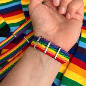 May include: A rainbow-striped watch strap with a silver buckle is fastened on a wrist. The strap's colours include red, orange, yellow, green, blue, and purple. Rainbow-coloured socks are in the background.