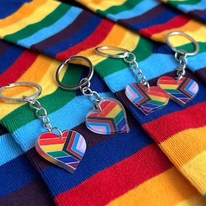 Op de afbeelding: Sleutelhangers met hartvormige hangers in een regenboogontwerp. De sleutelhangers hebben zilverkleurige ringen en kettingen. De achtergrond is een kleurrijk gestreept patroon in regenboogkleuren.