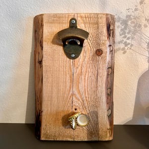 Puede incluir: Un abridor de botellas de madera rústico con un abridor de botellas de latón unido a él. Hay dos tapas de botellas doradas unidas a la madera.