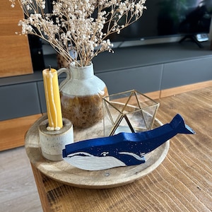 Könnte beinhalten: Eine blaue und weiße bemalte Holz-Walfigur, die auf einem Holztablett mit einem Kerzenhalter und einer Vase mit getrockneten weißen Blumen steht.