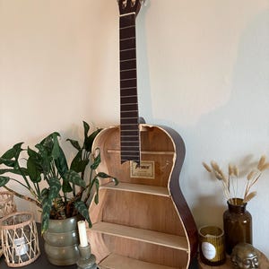 Può includere: Un corpo di chitarra acustica riutilizzato come scaffale da parete. L'esterno in legno marrone scuro presenta un interno svuotato con tre ripiani orizzontali in legno chiaro e un'etichetta del marchio Prince all'interno.