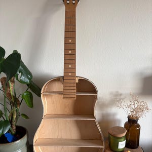 Könnte beinhalten: Ein gitarrenförmiges Regal aus hellem Holz mit drei Regalböden. Das Gitarrenregal hat eine natürliche Oberfläche. Hals und Kopfplatte der Gitarre sind sichtbar. Das Regal wird vor einer weißen Wand präsentiert.
